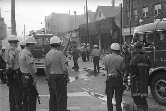 Cleveland police, wearing riot helmets and some armed with shotguns, protect firemen working on Hough Avenue in Cleveland, July 23, 1966. Police protection was ordered after three persons were arrested for heckling and abusing firemen. Several buildings were burned last night when rioting swept the predominantly black east side area. (AP Photo/Julian C. Wilson)